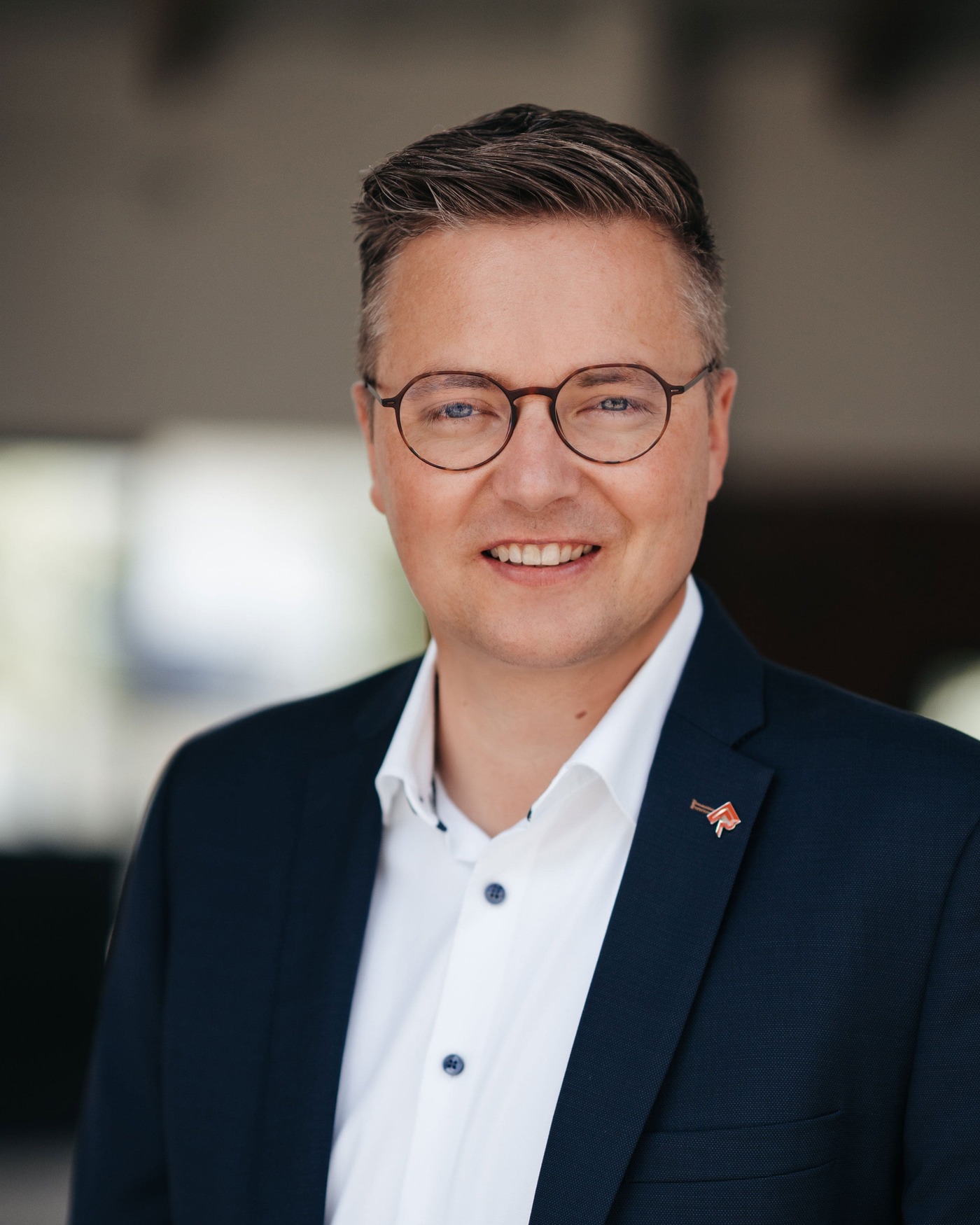 Ein Mann in einem professionellen Anzug lächelt freundlich. Im Hintergrund ist eine teils beleuchtete Reithalle zu erkennen. Die Atmosphäre ist modern und einladend, typisch für Viebrock Reithallen-Designs.