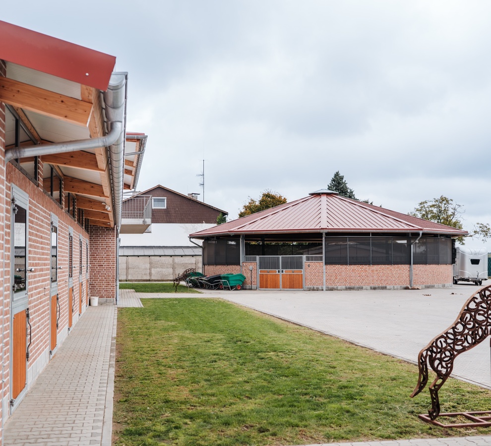 Stallgebäude aus rotem Klinker mit Holztüren an den Außenboxen, Rasenfläche mit Pferdeskulptur und im Hintergrund die Longierhalle