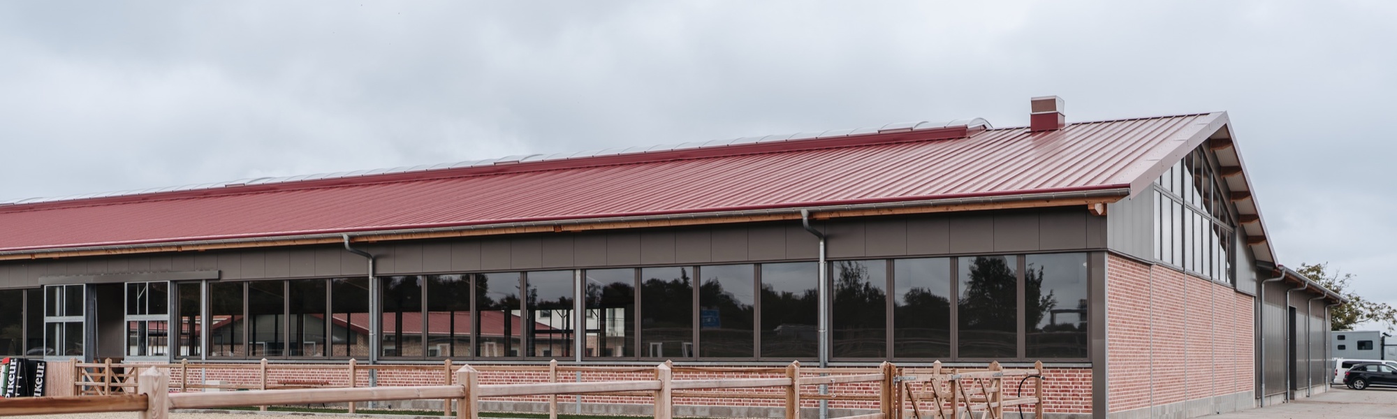 Reithalle mit rotem Dach, Klinkersockel und durchgehendem Fensterband, davor ein Reitplatz mit Holzzaun