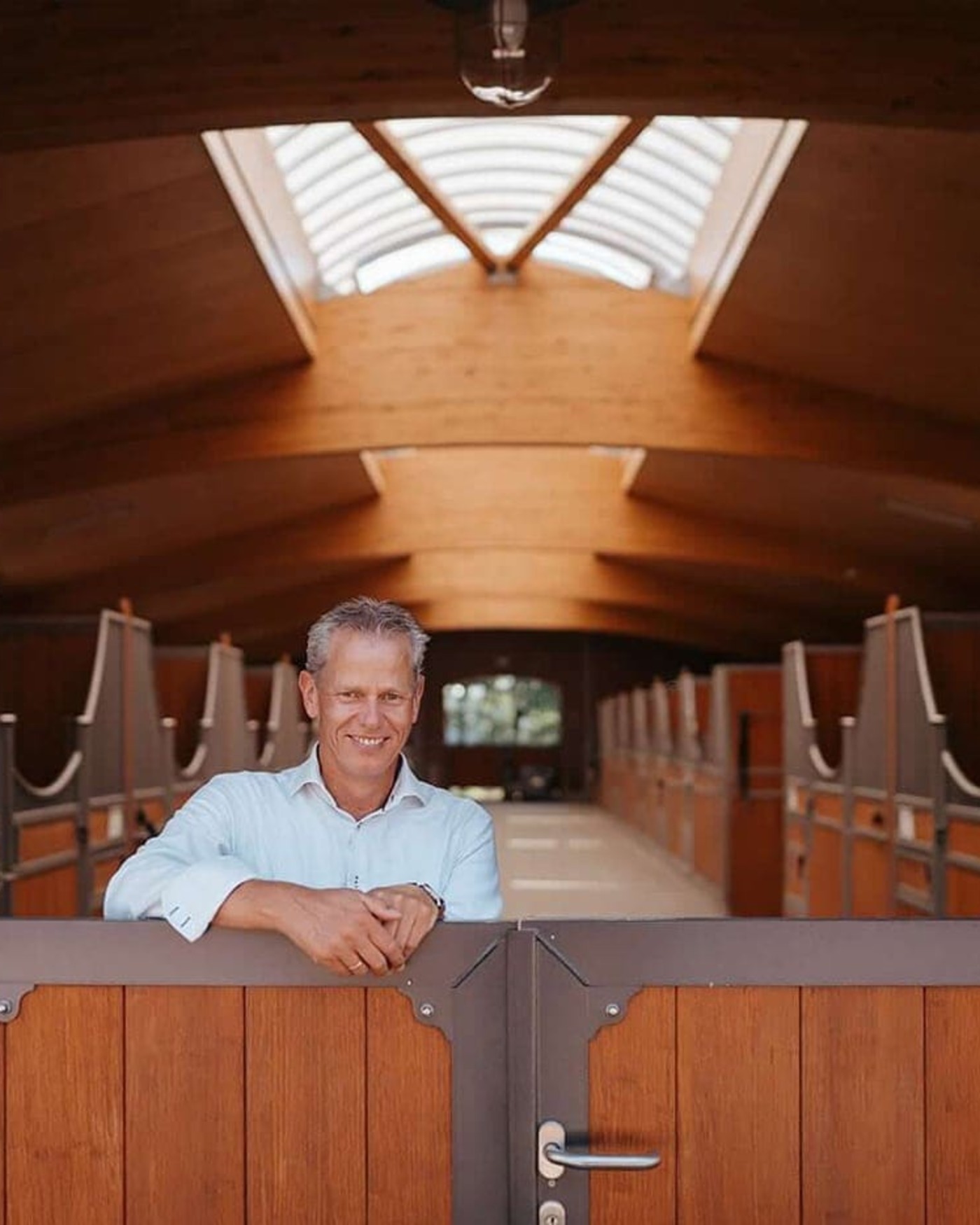 Ein Viebrock Reithallen Stall mit hochwertigen Holzställen und einem modernen, hellen Innenraum. Torsten Wessels steht lächelnd am Tor, umgeben von eleganten Boxen und einer großzügigen Dachkonstruktion mit Oberlichtern, die viel Tageslicht hereinlässt.