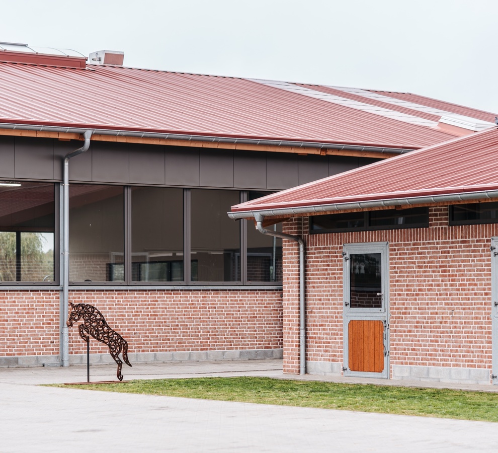 Reithalle und Stallgebäude aus rotem Klinker mit roten Dächern, davor die Metallskulptur eines springenden Pferdes